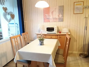 Zona pranzo con tavolo ricoperto da una tovaglia bianca, sedie di legno, una pianta in vaso, un forno a microonde, un tostapane e una lampada a sospensione.