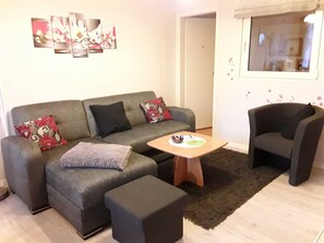En stue med en grå sofa, to lænestole, et lille træbord og en mørk måtte.