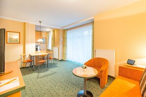 Et hotelværelse med spisebord, stole, sofa, et lille bord med en vase og blomster, et fjernsyn samt et vindue med gardiner.