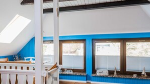 A room with a sloped ceiling, a wooden floor, a white railing, and a window with a view of the outside. - Beach whispers in the attic - Strandgeflüster (Ahrenshoop)