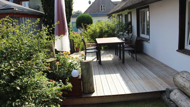 A wooden deck with a table and chairs, surrounded by greenery and a thatched-roof building. - Beach whispers in the attic - Strandgeflüster (Ahrenshoop)