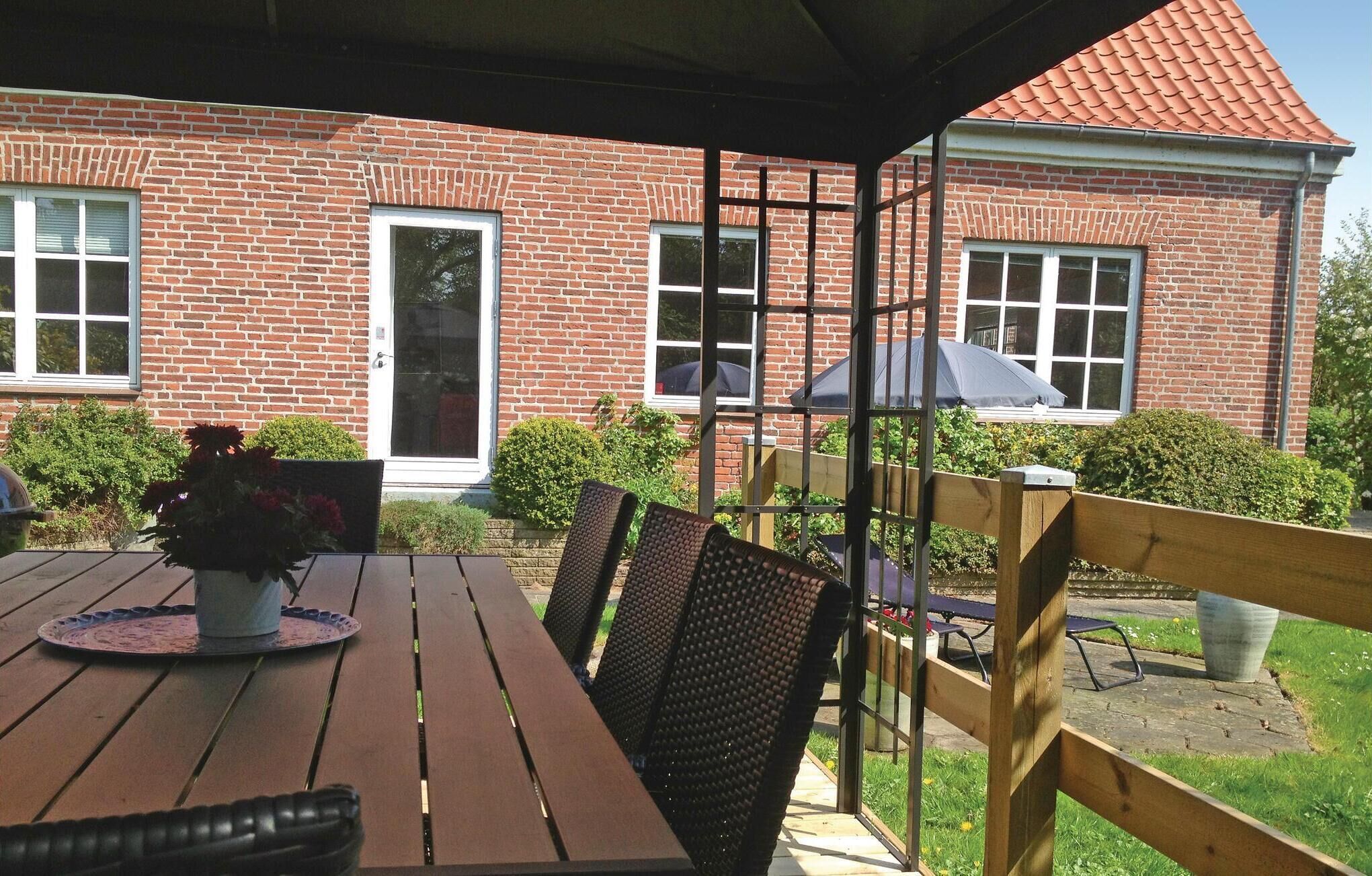 Une terrasse couverte avec une table et des chaises en bois, une plante en pot et, au fond, un bâtiment en briques doté de fenêtres et d’une porte.