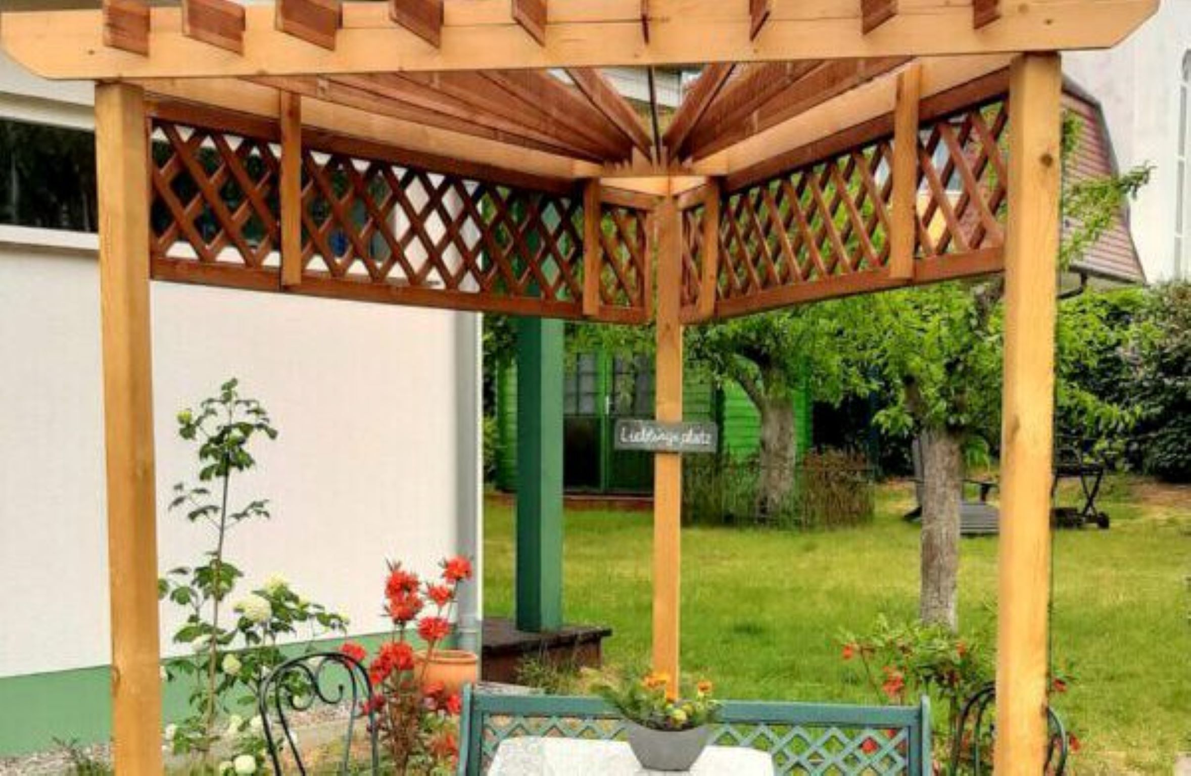 A wooden pergola structure with lattice panels, a garden with various plants and flowers, and a sign that reads "Liebesgarten".