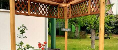 A wooden pergola structure with lattice panels, a garden with various plants and flowers, and a sign that reads "Liebesgarten".