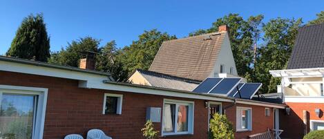 A brick house with solar panels, white plastic chairs, and a potted plant.