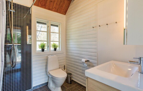Un baño con inodoro blanco, lavamanos, ventana con plantas y zona de ducha.
