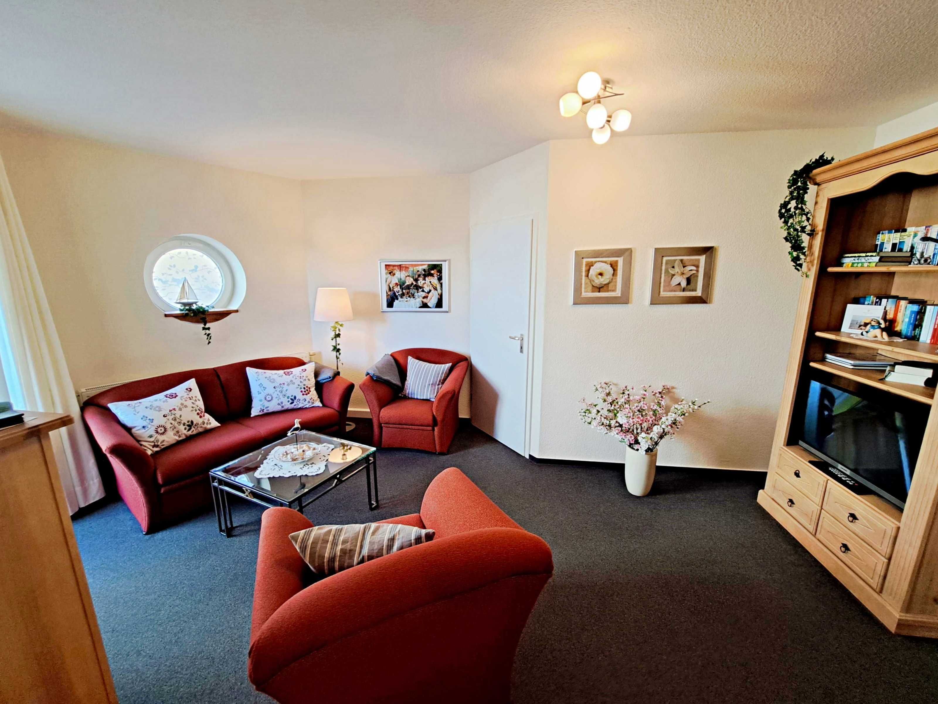Una sala de estar con un sofá rojo, un sillón rojo más pequeño, una mesa de centro, un mueble de TV con televisor de pantalla plana y una lámpara de pared.