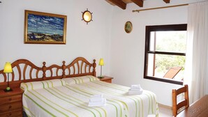 A bedroom with a bed, a nightstand, a lamp, a painting, a window, and a view of greenery outside.