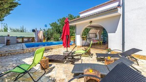 Zona junto a la piscina con sillones de descanso, una sombrilla roja y vistas a un edificio con tejado de tejas.