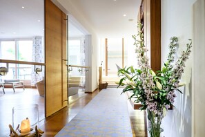 Un pasillo de hotel con suelo de madera, una puerta de cristal que da a una zona de estar y un jarrón con flores sobre una mesa.