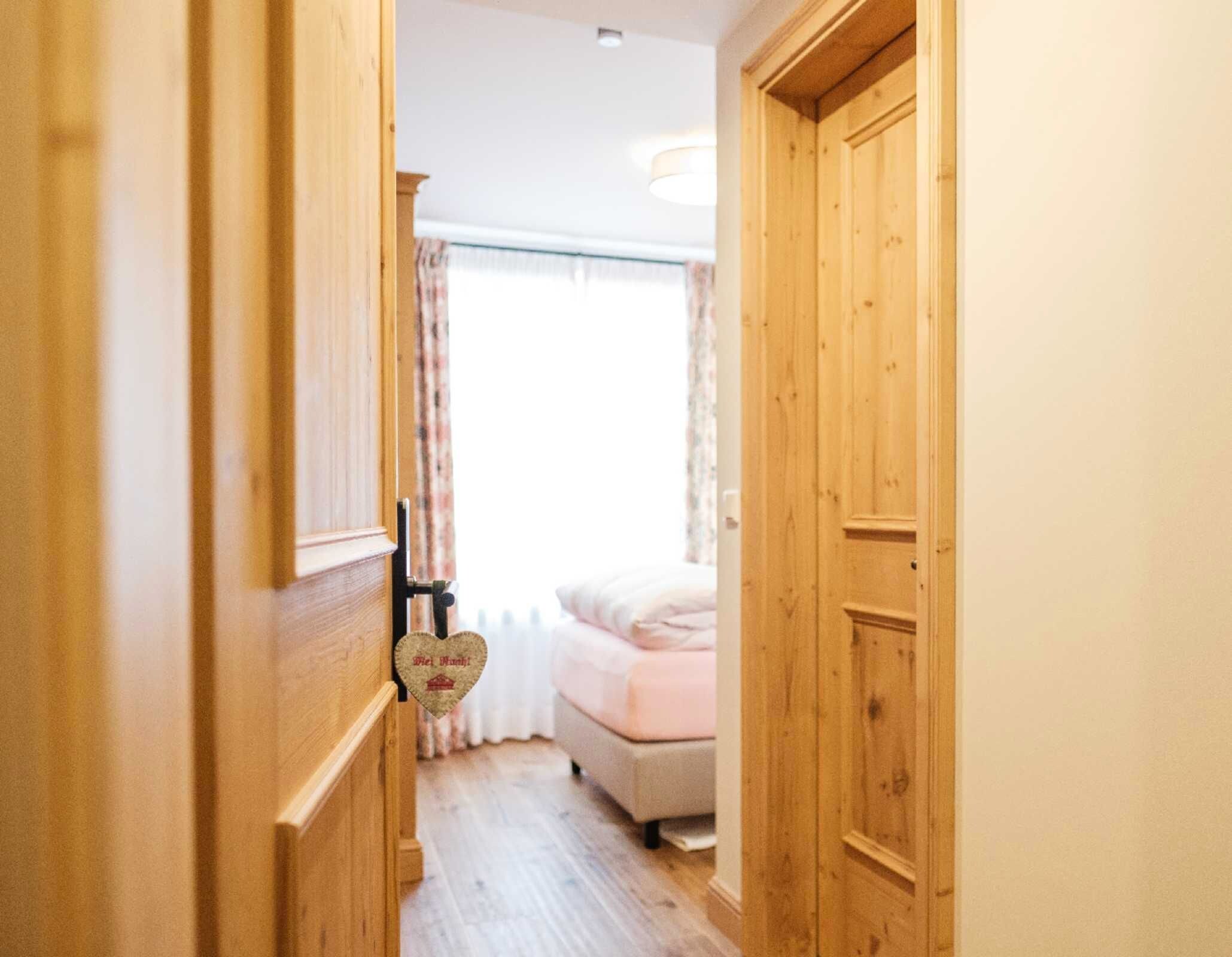 Un couloir étroit avec des portes en bois, un lit doté d’une tête de lit blanche et une fenêtre avec des rideaux.