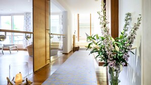 A hotel hallway with a wooden floor, a glass door leading to a seating area, and a vase with flowers on a table. - Single Room - Landhaus am Stein (Bad Wiessee)