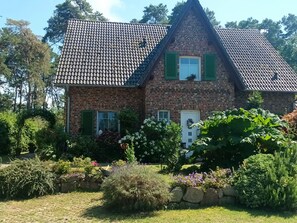 A two-story stone house with a tiled roof, green shutters, and a garden with various plants and flowers. - Lod - Goldbach - Holiday apartment (Loddin)