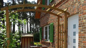A brick building with a wooden pergola, a table with chairs, and a window with green shutters. - Lod - Goldbach - Holiday apartment (Loddin)
