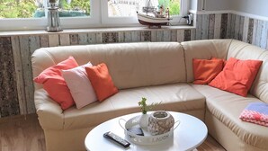 A beige sectional sofa with orange and pink cushions, a white coffee table with a remote control and decorative items, and a window with a view of greenery and a model ship. - Lod - Goldbach - Holiday apartment (Loddin)