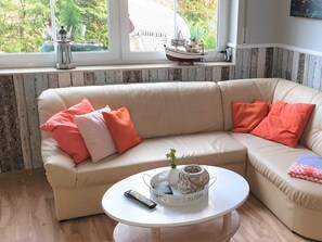 Un sofá seccional color beige con cojines naranja y rosa, una mesa de centro blanca con un control remoto y objetos decorativos, y una ventana con vistas a la vegetación y un barco modelo.
