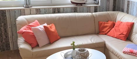 A beige sectional sofa with orange and pink cushions, a white coffee table with a remote control and decorative items, and a window with a view of greenery and a model ship.