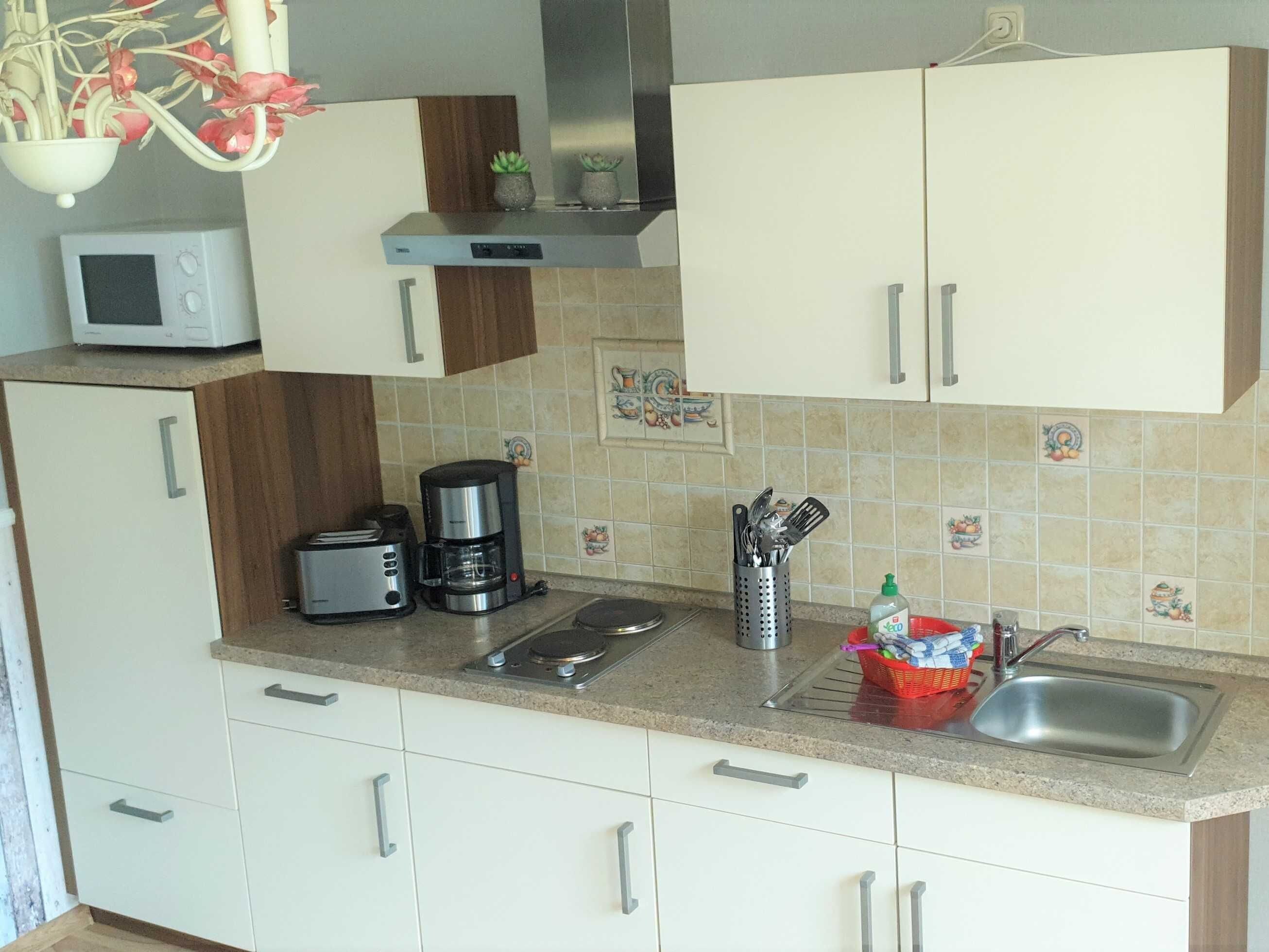 A compact kitchen with a microwave, toaster, coffee maker, and a sink with a red basket of utensils.