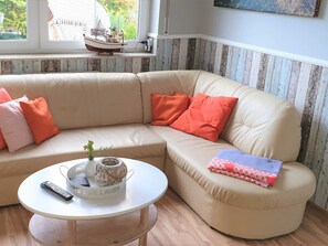 A beige sectional sofa with orange cushions, a white coffee table with a remote control and decorative items, and a window with a view of greenery and a model ship. - Lod - Goldbach - Holiday apartment (Loddin)