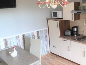 Une cuisine équipée d’un four à micro-ondes, d’un grille-pain et d’une machine à café sur le plan de travail, d’un meuble blanc et d’une statuette de poulet décorative posée sur la table.