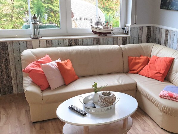 Un canapé d’angle beige agrémenté de coussins orange et roses, une table basse blanche avec une télécommande et des objets décoratifs, et une fenêtre donnant sur un paysage verdoyant et un modèle de bateau.