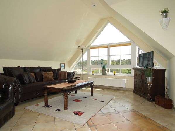 A spacious loft with a high ceiling, a large window offering a view of the outdoors, a dark leather sofa, a wooden coffee table, and a television on a wooden cabinet.