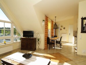 A spacious living area with a television on a wooden cabinet, a dining table with chairs, and a view of the outdoors through large windows.