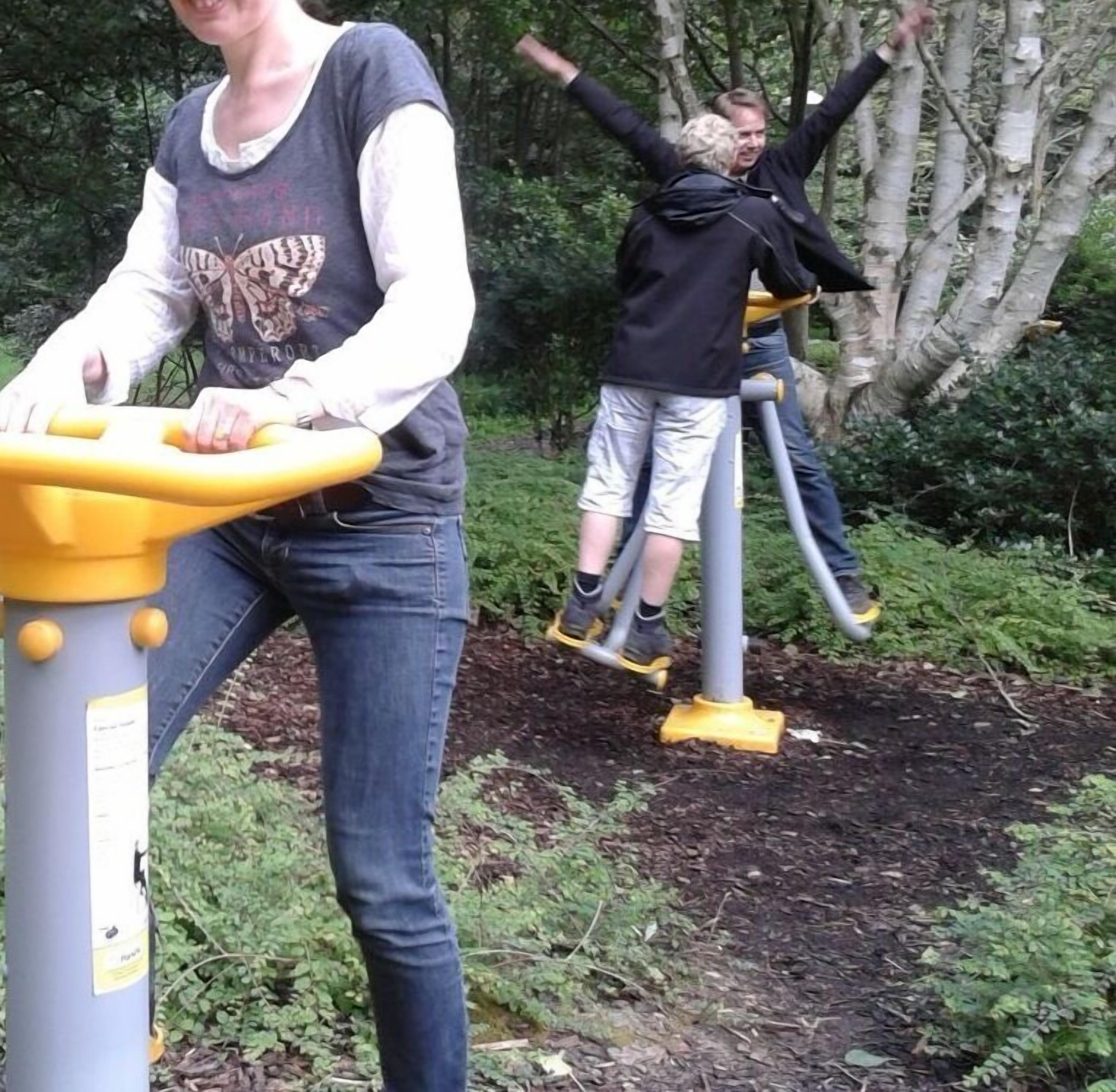 Dos personas usando equipo de ejercicio al aire libre en un parque con árboles y follaje de fondo.