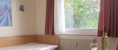Un dormitorio pequeño y ordenado con una cama individual, una mesa con una botella y un vaso, y una ventana con cortinas.