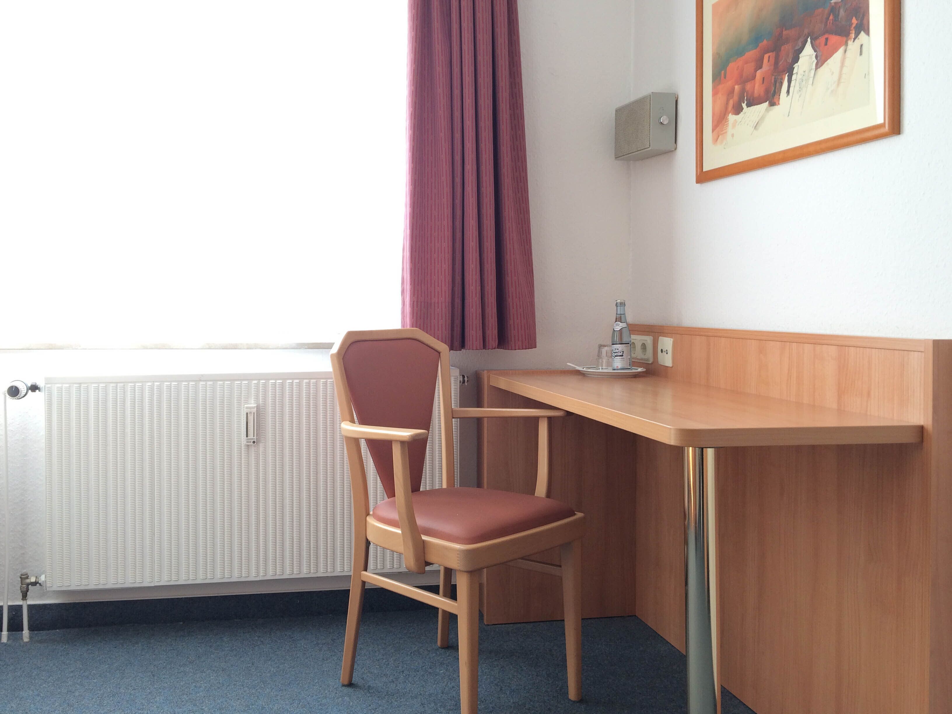 A hotel room with a wooden desk, a chair, a radiator, a window with curtains, and a painting on the wall.