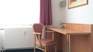 A hotel room with a wooden desk, a chair, a radiator, a window with curtains, and a painting on the wall.