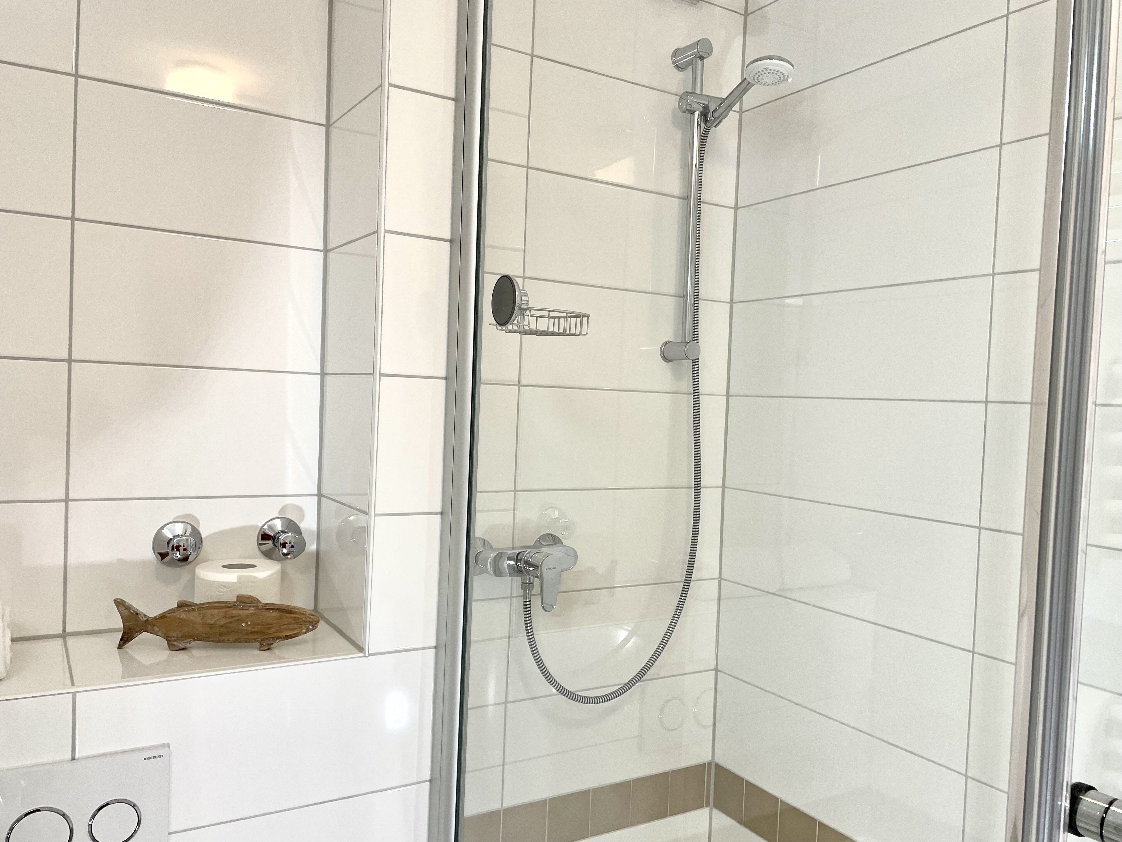 Un baño moderno con ducha compartimentada por vidrio, azulejos blancos y un estante con artículos de aseo.