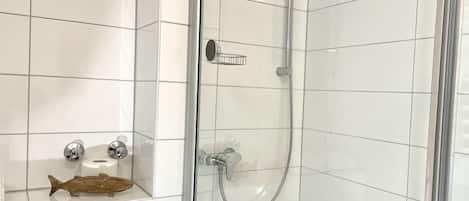 Un baño moderno con ducha compartimentada por vidrio, azulejos blancos y un estante con artículos de aseo.