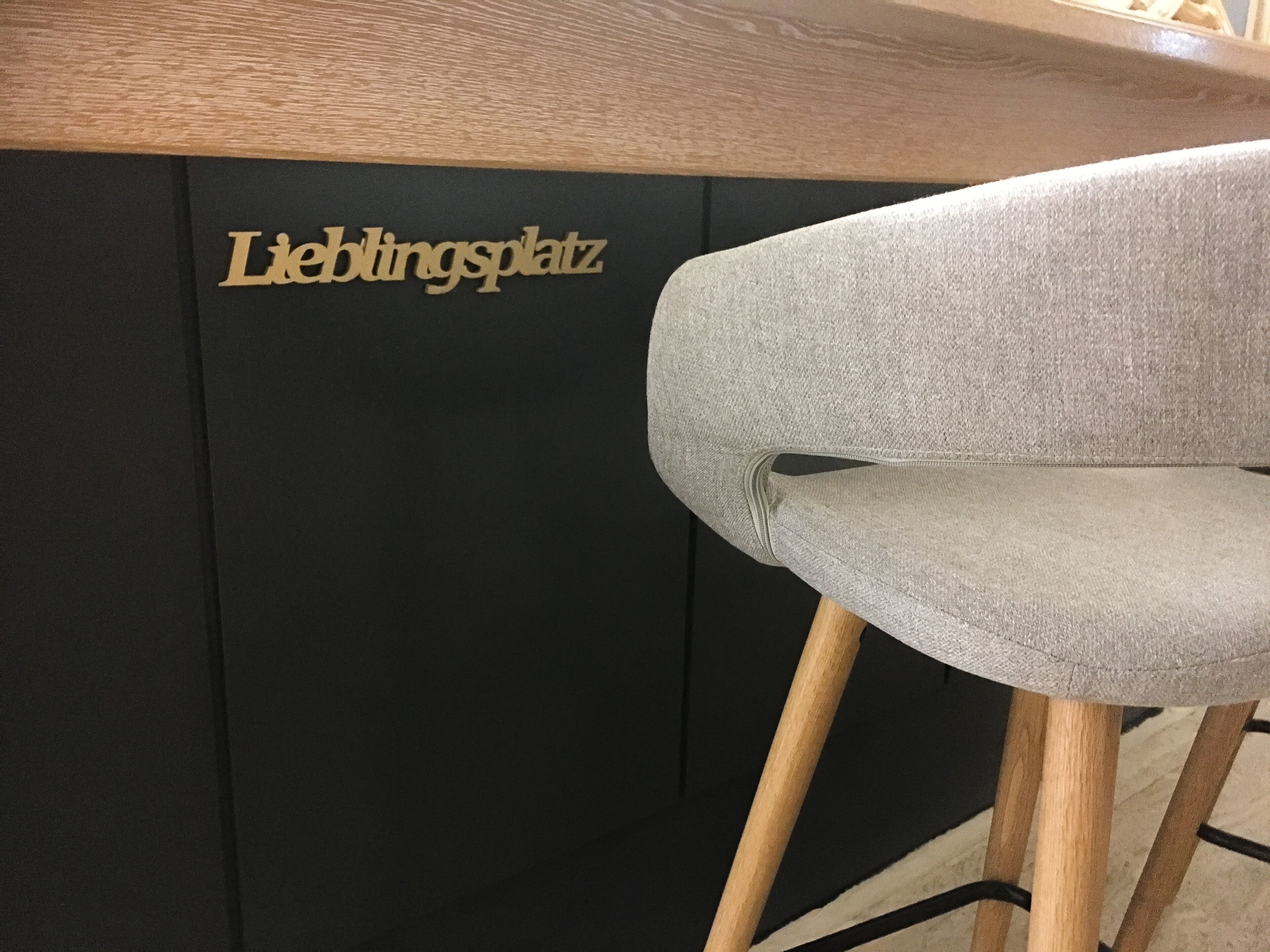 Un taburete con asiento tapizado gris y patas de madera, situado frente a una pared negra con el texto 