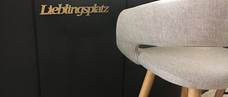 Un taburete con asiento tapizado gris y patas de madera, situado frente a una pared negra con el texto
