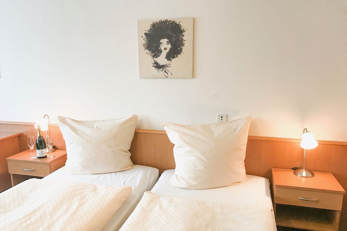 A hotel room with two beds, white linens, a nightstand with a bottle of champagne and two glasses, a wall-mounted artwork, and a bedside lamp.