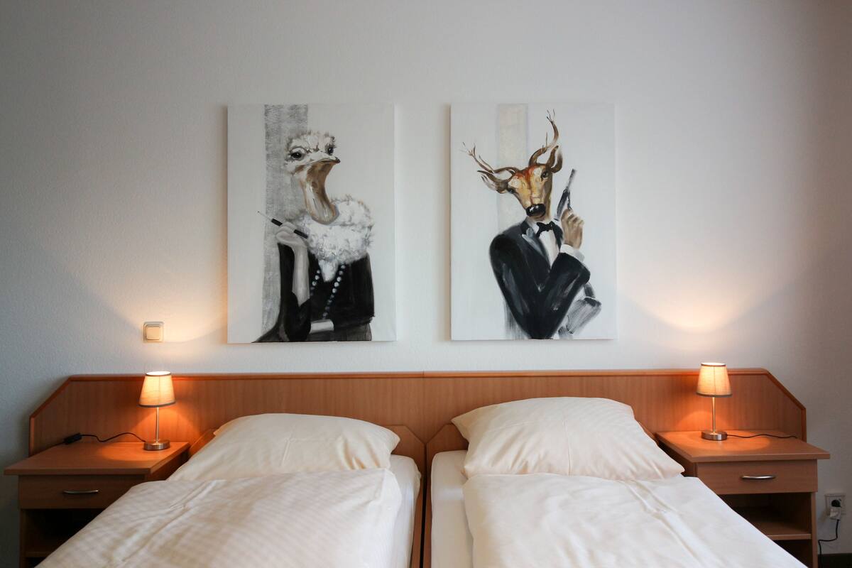 A hotel room with two beds, each with a bedside table and lamp. There are two framed artworks on the wall above the beds.