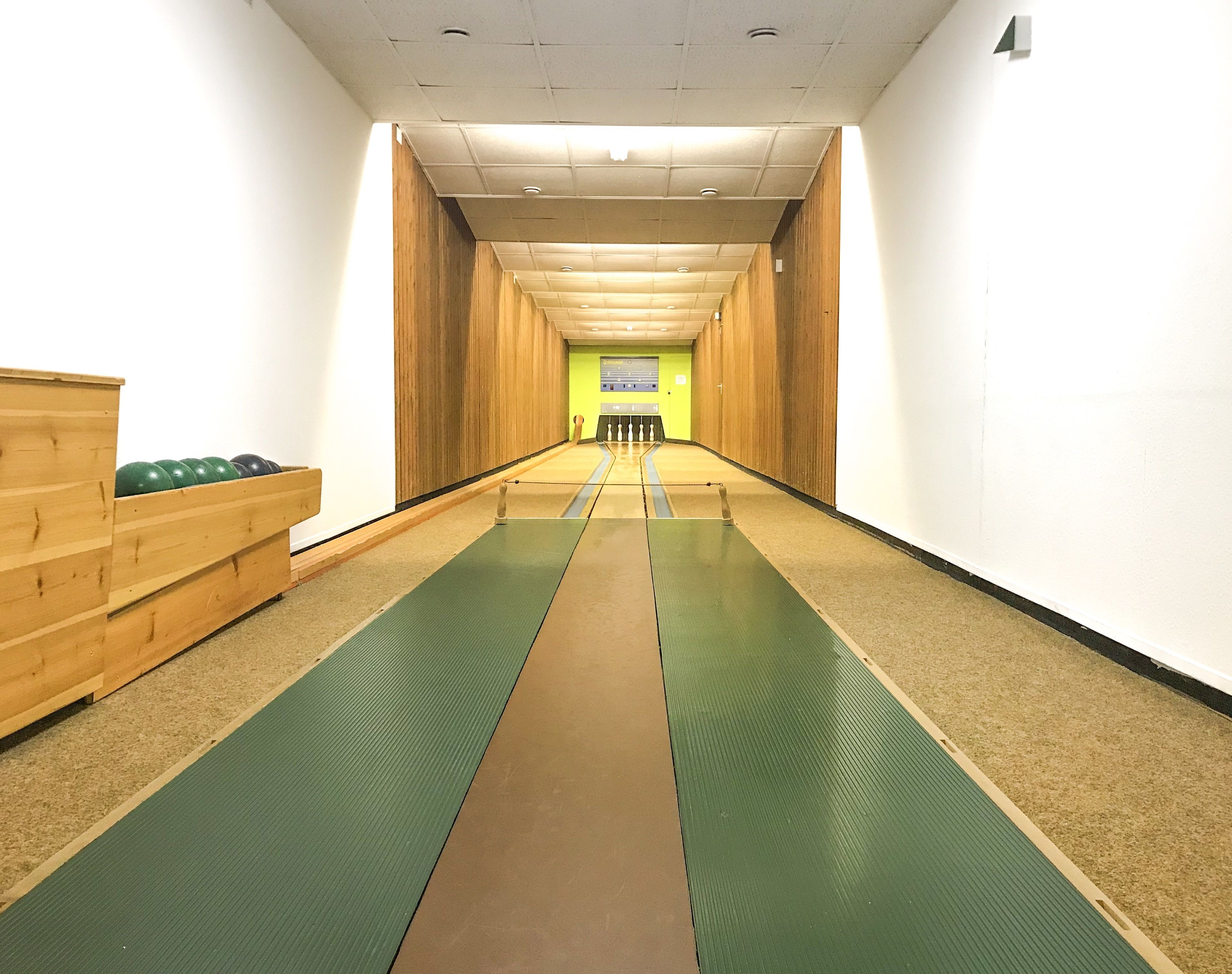 Un pasillo con un banco de madera a la izquierda, suelo verde y marrón, y pared de madera a la derecha.