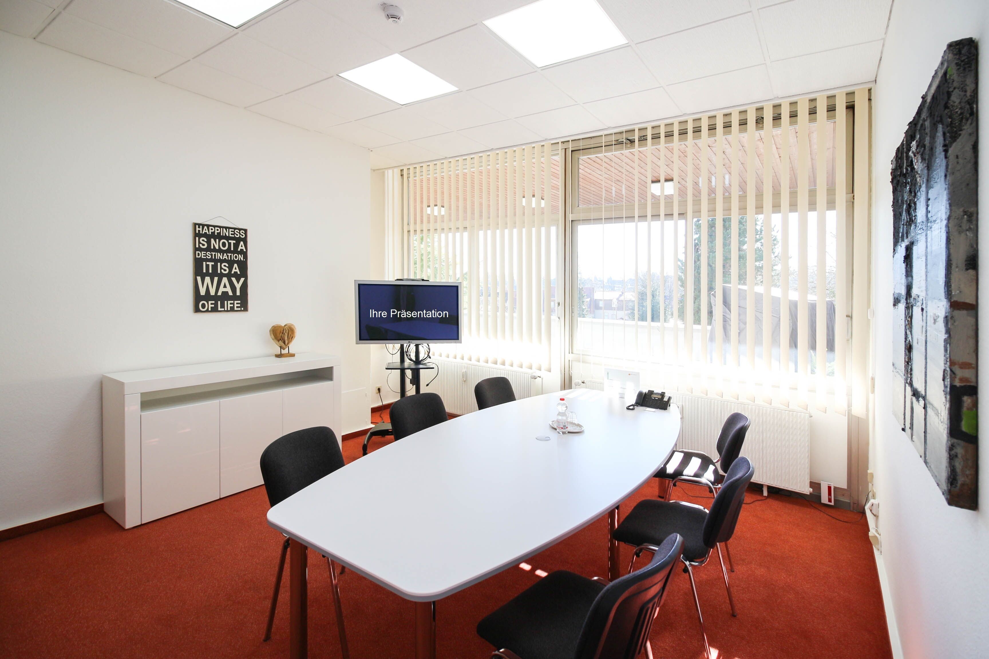 A conference room with a white table, black chairs, a TV, and a large window with blinds.