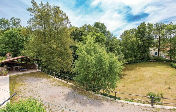 A rustic outdoor area with a stone barbecue and seating, surrounded by dense trees and a grassy field. - Gorgeous home in Poljane with sauna (Poljane)