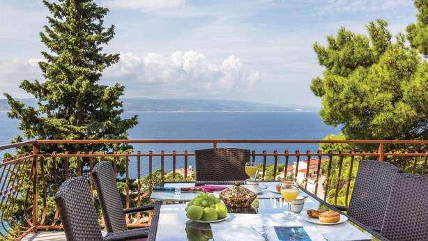 A balcony with wicker furniture, a table with fruits, a glass of juice, and a view of the sea and mountains. - 4 bedroom beautiful home in Brela (Brela)