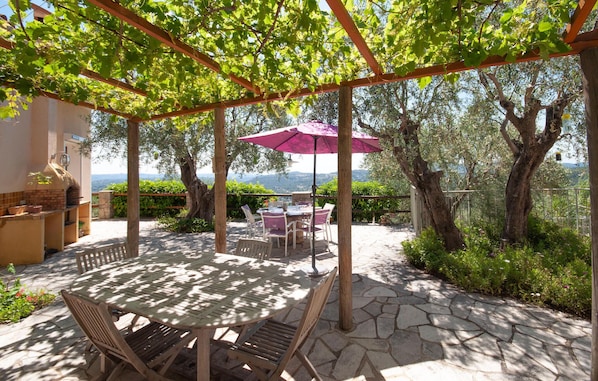 An outdoor patio with a stone table and chairs, a pergola with a purple umbrella, and a view of the landscape.
