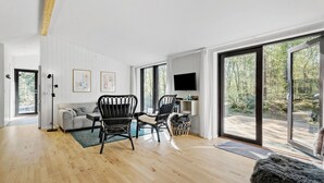 Un vaste salon doté d’un plancher en bois, d’un canapé gris, de fauteuils en osier noir, d’un téléviseur fixé au mur et de grandes portes-fenêtres coulissantes donnant sur une zone boisée.