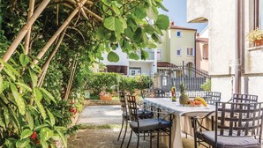 Un patio al aire libre con mesa y sillas, rodeado de exuberante follaje y vistas a un edificio con tejado de baldosas.