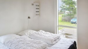 Une chambre avec une grande fenêtre, un lit avec une literie blanche et une prise électrique fixée au mur.