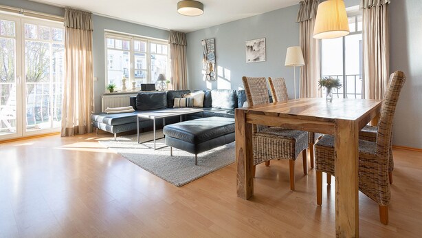 A spacious living room with a wooden dining table, wicker chairs, a blue sofa, and a large window with curtains. - Dünenstr. 3a Whg.Ki-01. - Ki-Whg. 01 (Kühlungsborn)