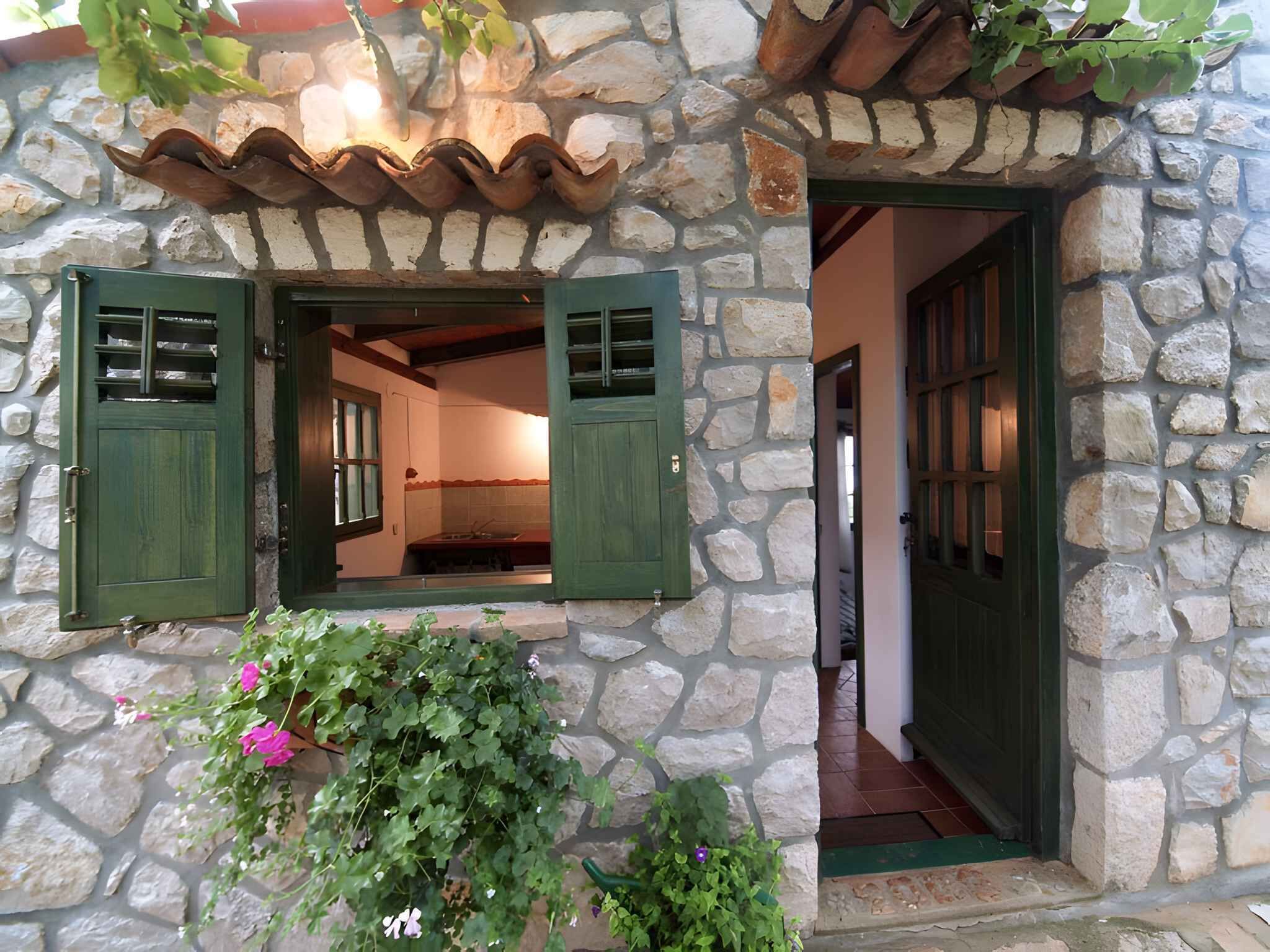 Un bâtiment en pierre avec une porte verte et des volets aux fenêtres, un toit en tuiles et un petit jardin fleuri.