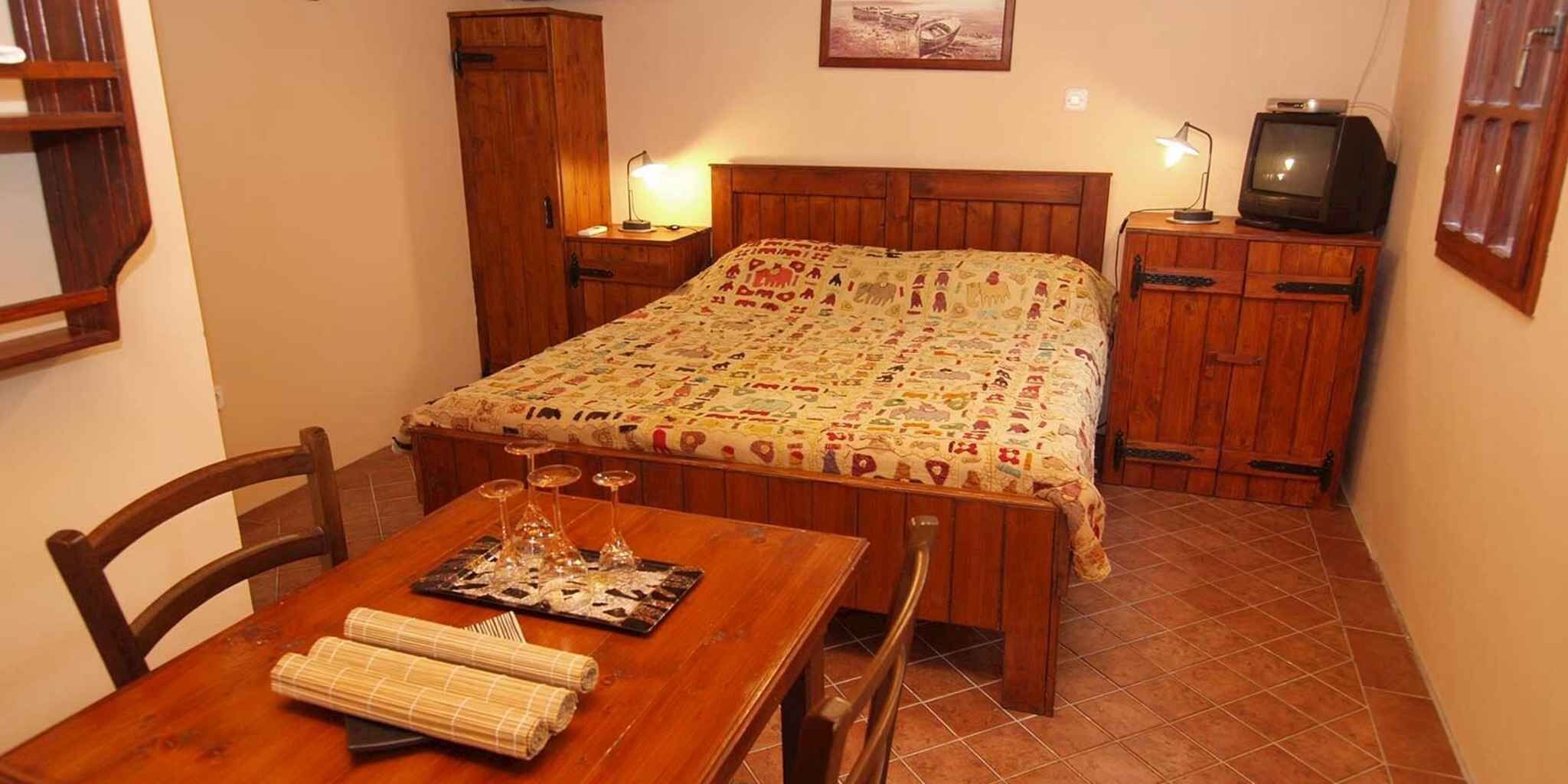 Ein kleiner, gemütlicher Raum mit einem hölzernen Bett, einem Tisch mit Gläsern und einer Speisekarte sowie einem Fernseher auf einer Holzkommode.