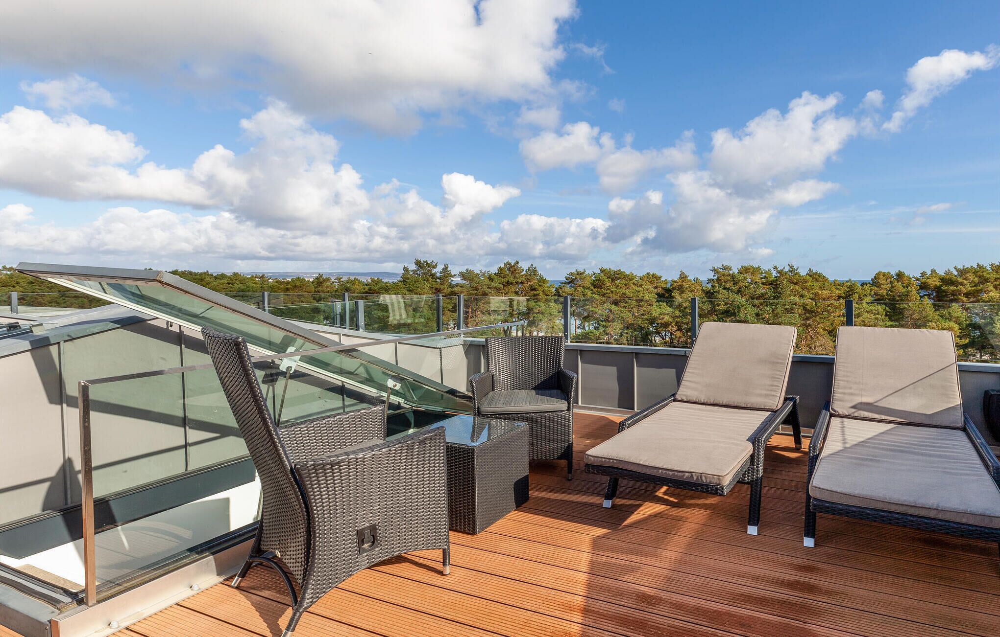 A rooftop terrace with glass balustrades, wicker furniture, and a view of the surrounding landscape.
