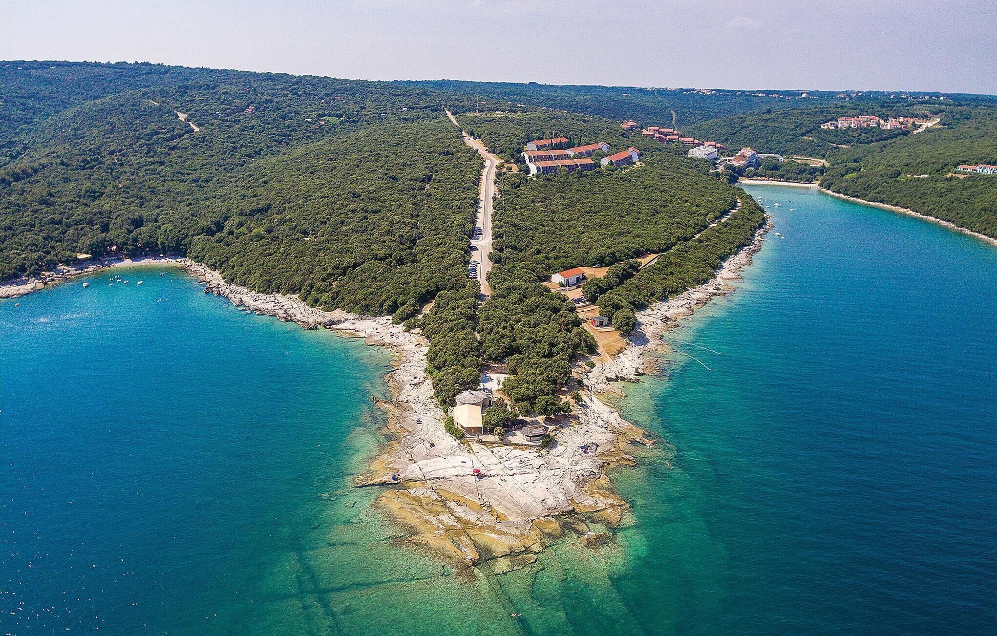 Vista aerea di una zona costiera con una stretta lingua di terra circondata dall'acqua su tre lati, con una strada, edifici e un mare blu cristallino.